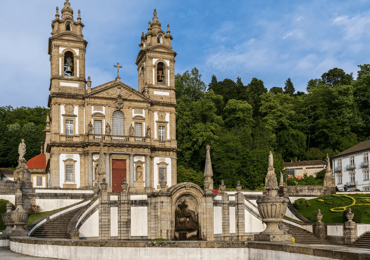 Fachada barroca do Santuário do Bom Jesus do Monte em Braga, com duas torres sineiras, escadaria monumental com estátuas e fonte, jardins floridos e vegetação exuberante ao fundo, em Portugal.