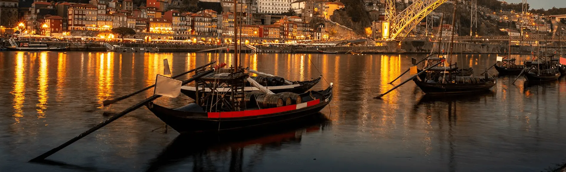 Barcos rabelos tradicionais com barris de vinho do Porto ancorados no Rio Douro à noite, com as luzes da Ribeira e a Ponte Dom Luís I refletidas na água ao fundo, em Portugal.