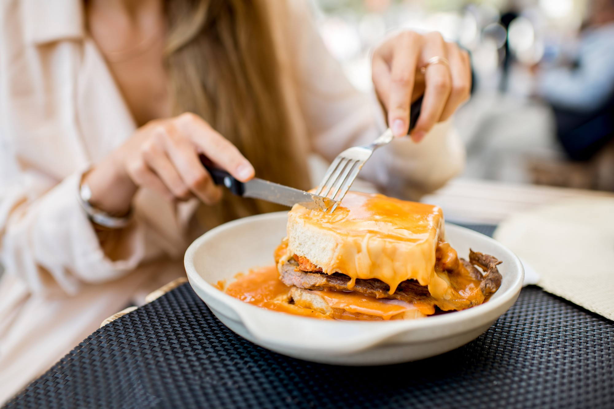 Mulher cortando uma francesinha com faca e garfo em restaurante, prato típico português coberto com queijo derretido e molho de tomate apimentado, servido em prato fundo branco.