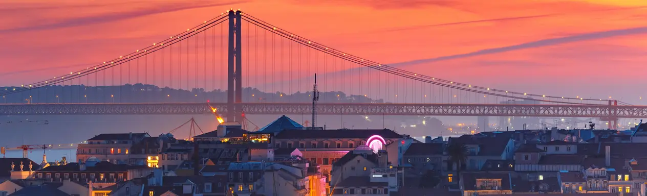 Vista panorâmica de Lisboa ao entardecer com a Ponte 25 de Abril iluminada sobre o Rio Tejo e o skyline da cidade em tons alaranjados e rosados, em Portugal.