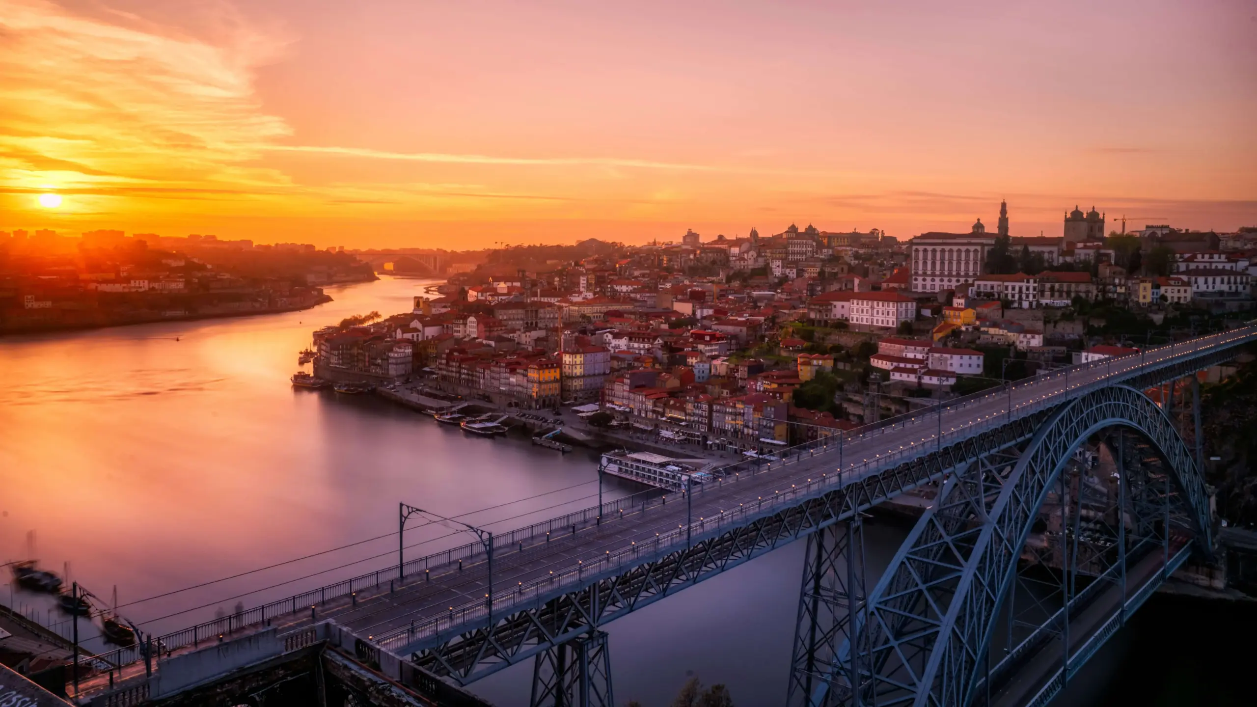 Vista aérea da Ponte Dom Luís I em primeiro plano ao pôr do sol, com o Rio Douro refletindo tons alaranjados e rosados, a Ribeira e o centro histórico do Porto iluminados ao fundo, em Portugal.