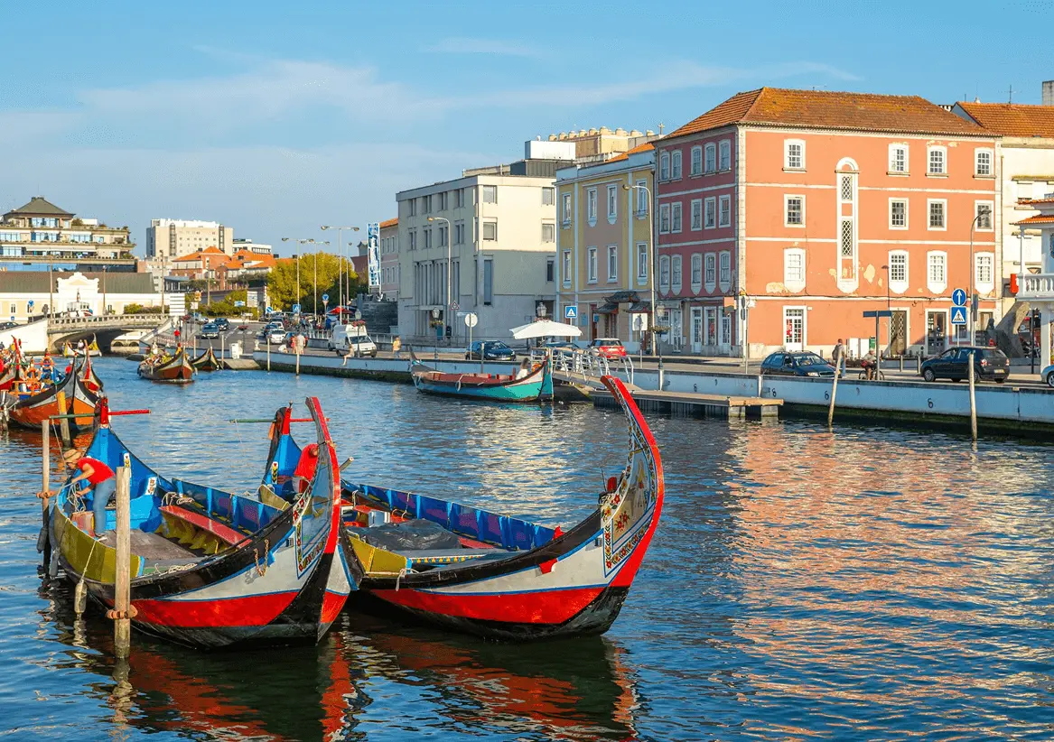 Barcos moliceiros coloridos atracados no canal central de Aveiro, com fachadas de edifícios históricos coloridos ao longo da margem e céu azul ao fundo, em Portugal.