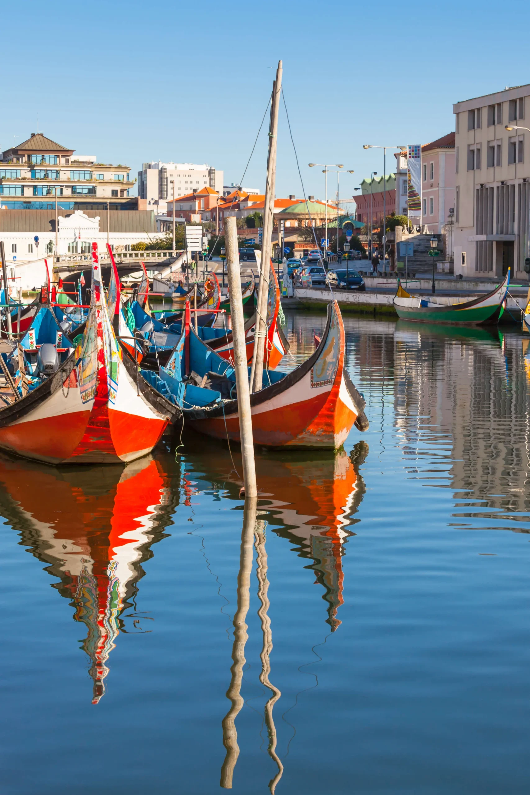Barcos moliceiros coloridos com proas laranja e azul atracados no canal de Aveiro, com reflexo nítido na água calma e edifícios da cidade ao fundo em dia de céu azul, em Portugal.