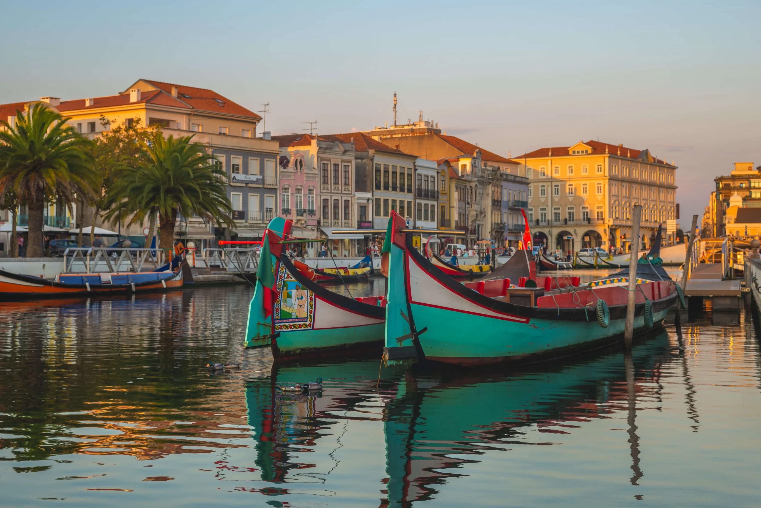 Barcos moliceiros coloridos atracados no canal central de Aveiro, com fachadas de edifícios históricos coloridos ao longo da margem e céu azul ao fundo, em Portugal.