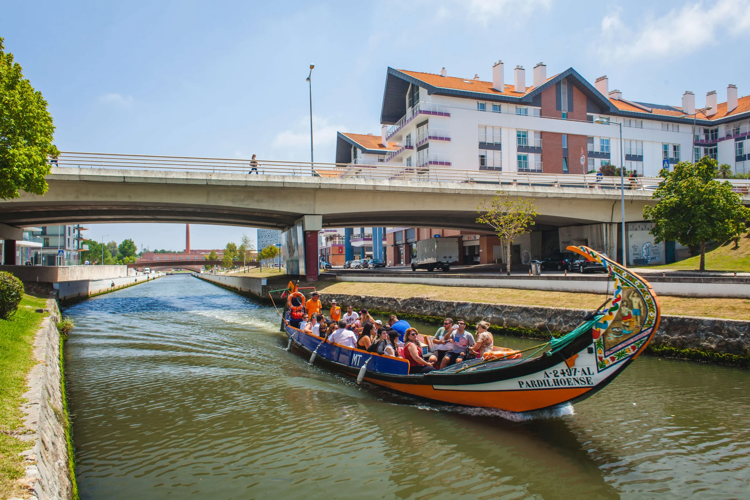 Grupo de turistas em passeio de barco moliceiro colorido navegando pelo canal de Aveiro em dia ensolarado, passando sob uma ponte com edifícios modernos ao fundo, em Portugal.