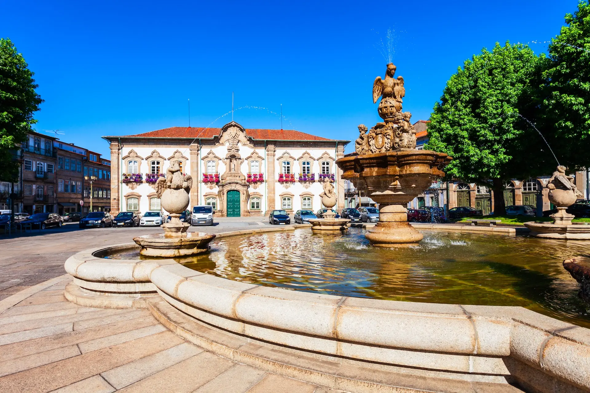 Fonte barroca com estátuas de galos em pedra na Praça do Município de Braga, com a fachada branca ornamentada da Câmara Municipal decorada com flores coloridas e céu azul ao fundo, em Portugal.