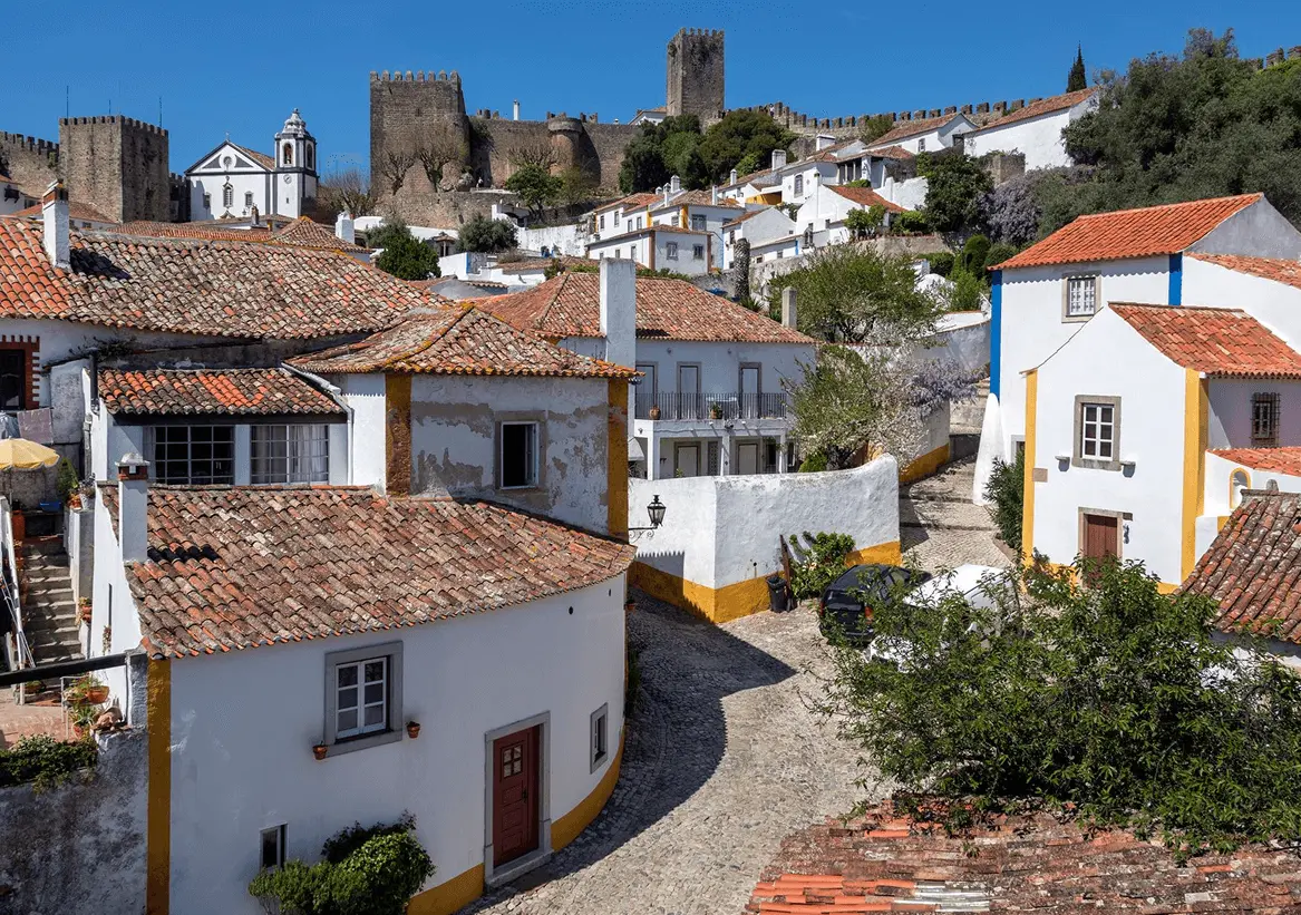 Viela de paralelepípedos em Óbidos com casas brancas de detalhes azuis e amarelos, arco de pedra medieval, lanterna de ferro forjado e colinas verdes ao fundo, em Portugal.