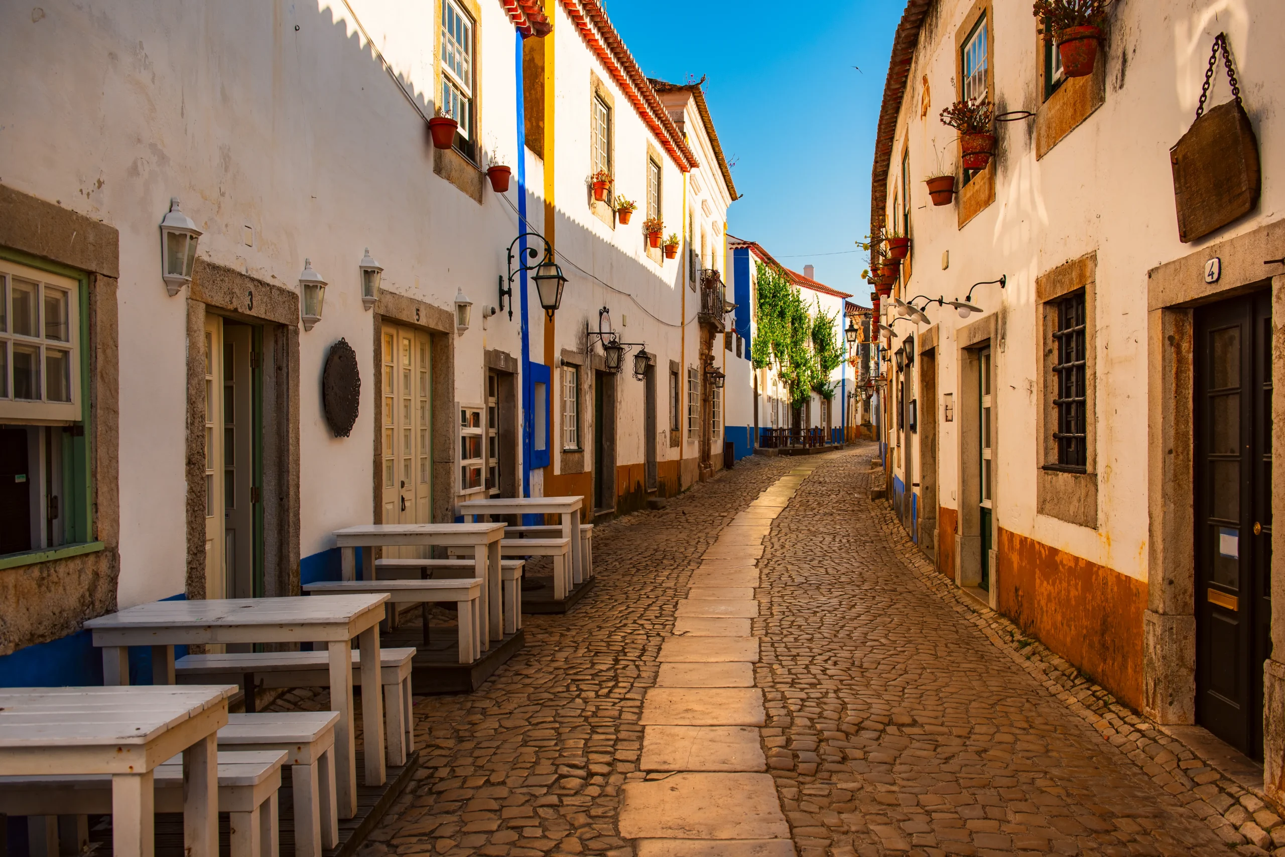 Rua de paralelepípedos vazia em Óbidos com casas brancas de detalhes coloridos, lanternas de ferro, vasos de flores nas fachadas, mesas de café ao ar livre e céu azul ao fundo, em Portugal.