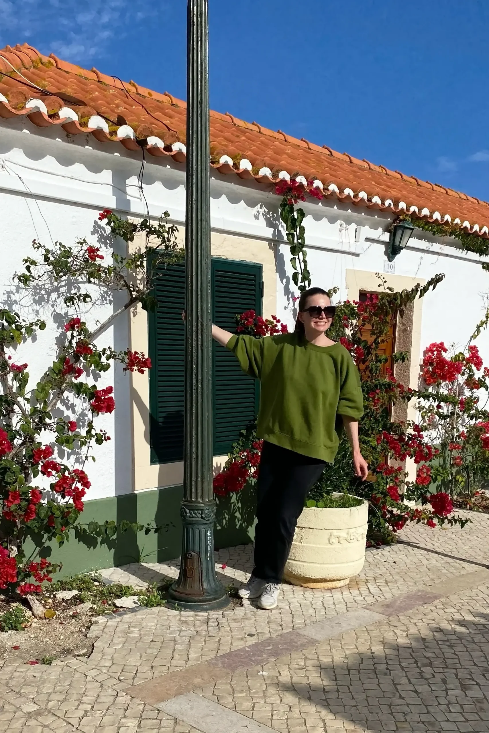 Turista sorrindo apoiada em poste de ferro forjado em rua de paralelepípedos de Óbidos, com casa branca coberta por buganvílias vermelhas e céu azul ao fundo, em Portugal.