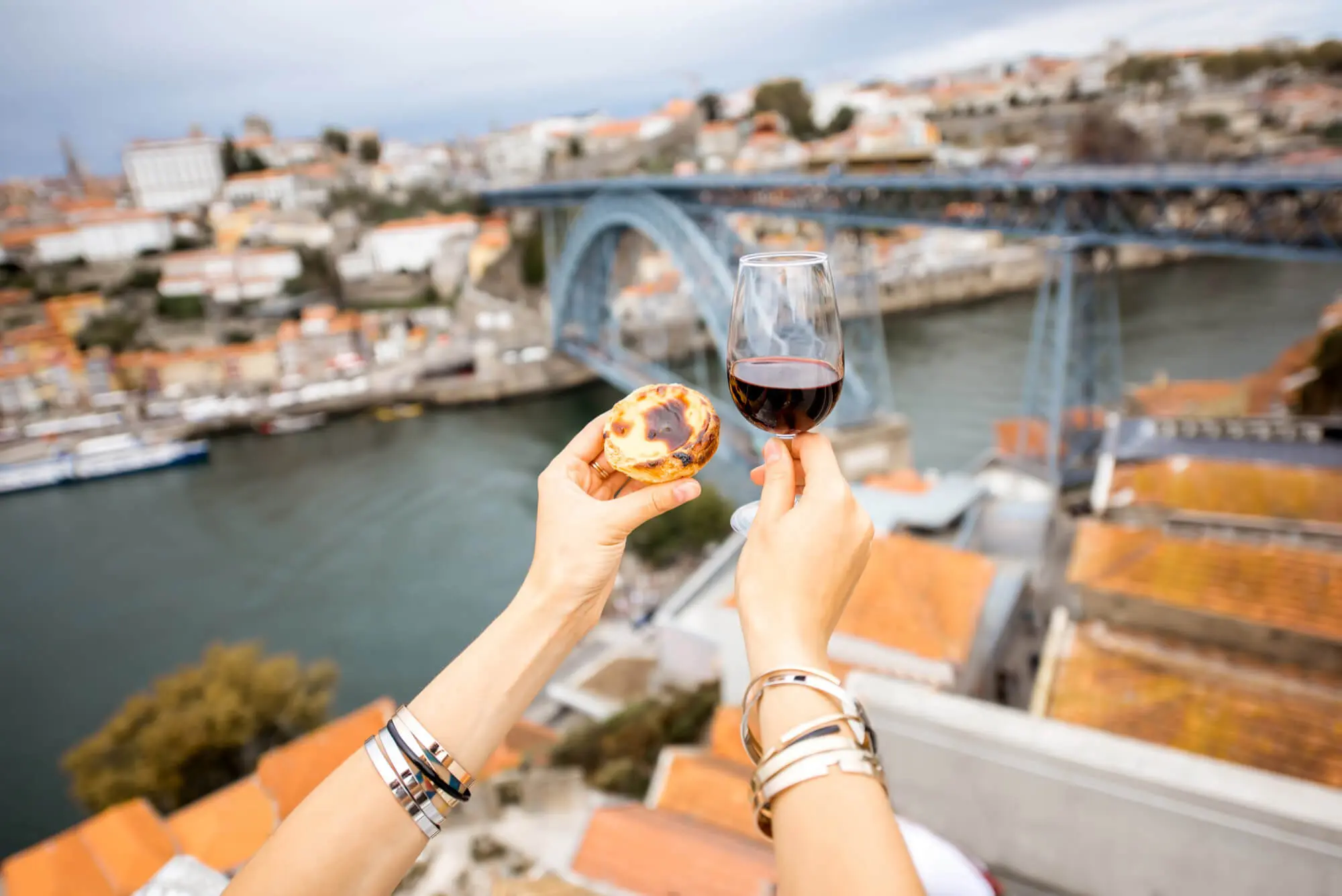 Mãos segurando um pastel de nata e uma taça de vinho do Porto com a Ponte Dom Luís I, o Rio Douro e o centro histórico do Porto desfocados ao fundo, em Portugal.